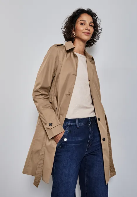 Frau mit beigem Trenchcoat, weißem Shirt und blauer Jeans vor grauem Hintergrund.
